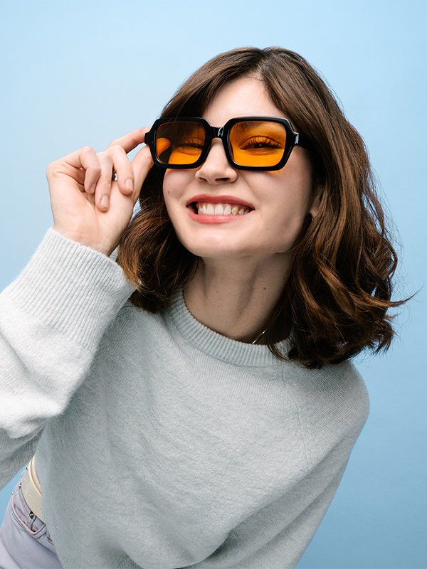 Eine junge Frau mit braunen Haaren und einer schwarzen Sonnenbrille mit orangen Gläsern.
