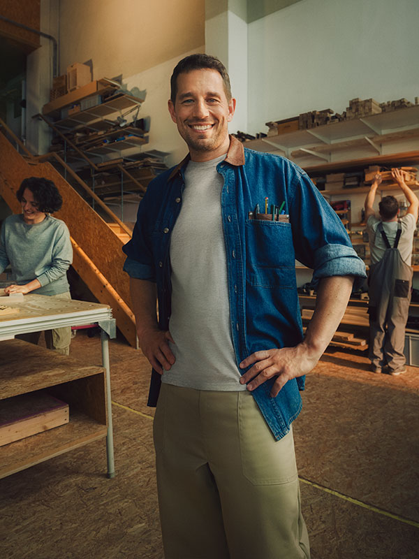 Ein Tischler steht in seiner Werkstatt. Her hat die Hände in die Hüften gestemmt und lächelt selbstbewusst in die Kamera.